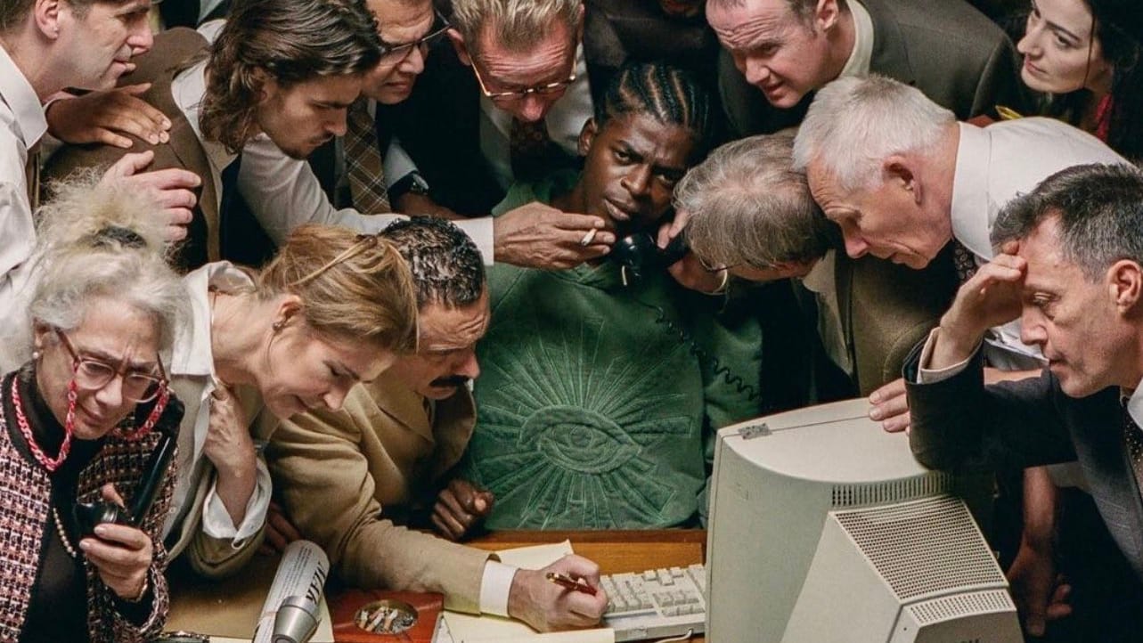 A man sitting at a desk. Many people surround him and do his tasks: holding his phone, holding his cigarette, etc.